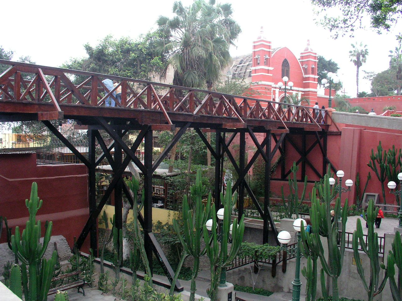 The wooden Puente de los Suspiros in Barranco, Lima, with the red brick shell of La Ermita church rising behind it under a grey sky