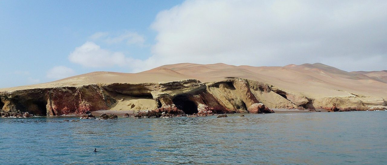 Red and ochre sea cliffs with desert dunes rising behind them above calm water on the Paracas coast near the Ballestas Islands