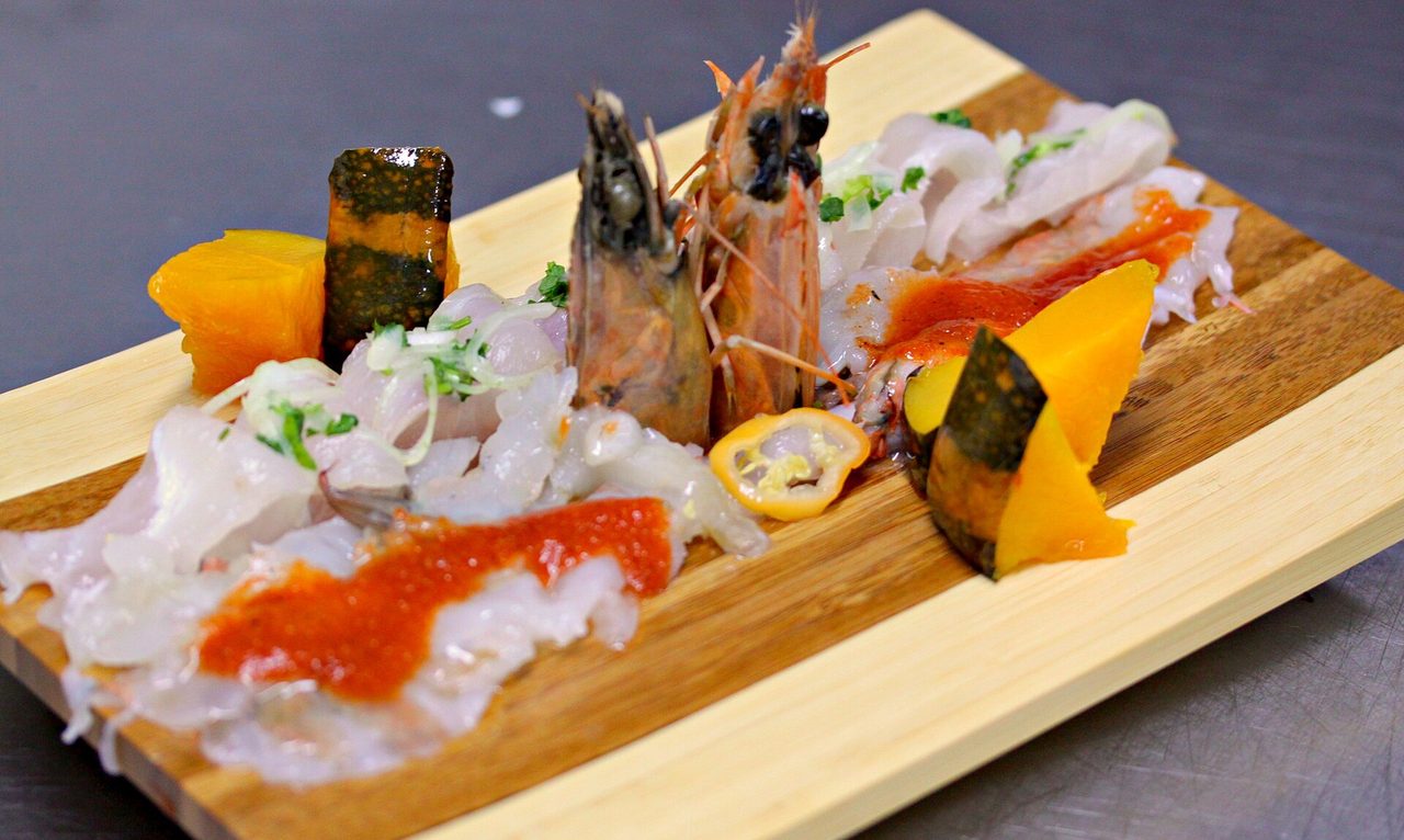 Two tiraditos on a wooden board: thin slices of raw white fish under red chilli sauce, with prawns and wedges of orange sweet potato