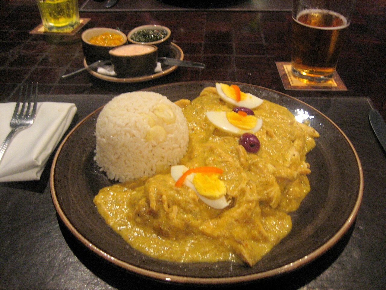 Aji de gallina served on a dark plate: shredded chicken in yellow chilli sauce with white rice, boiled egg and an olive