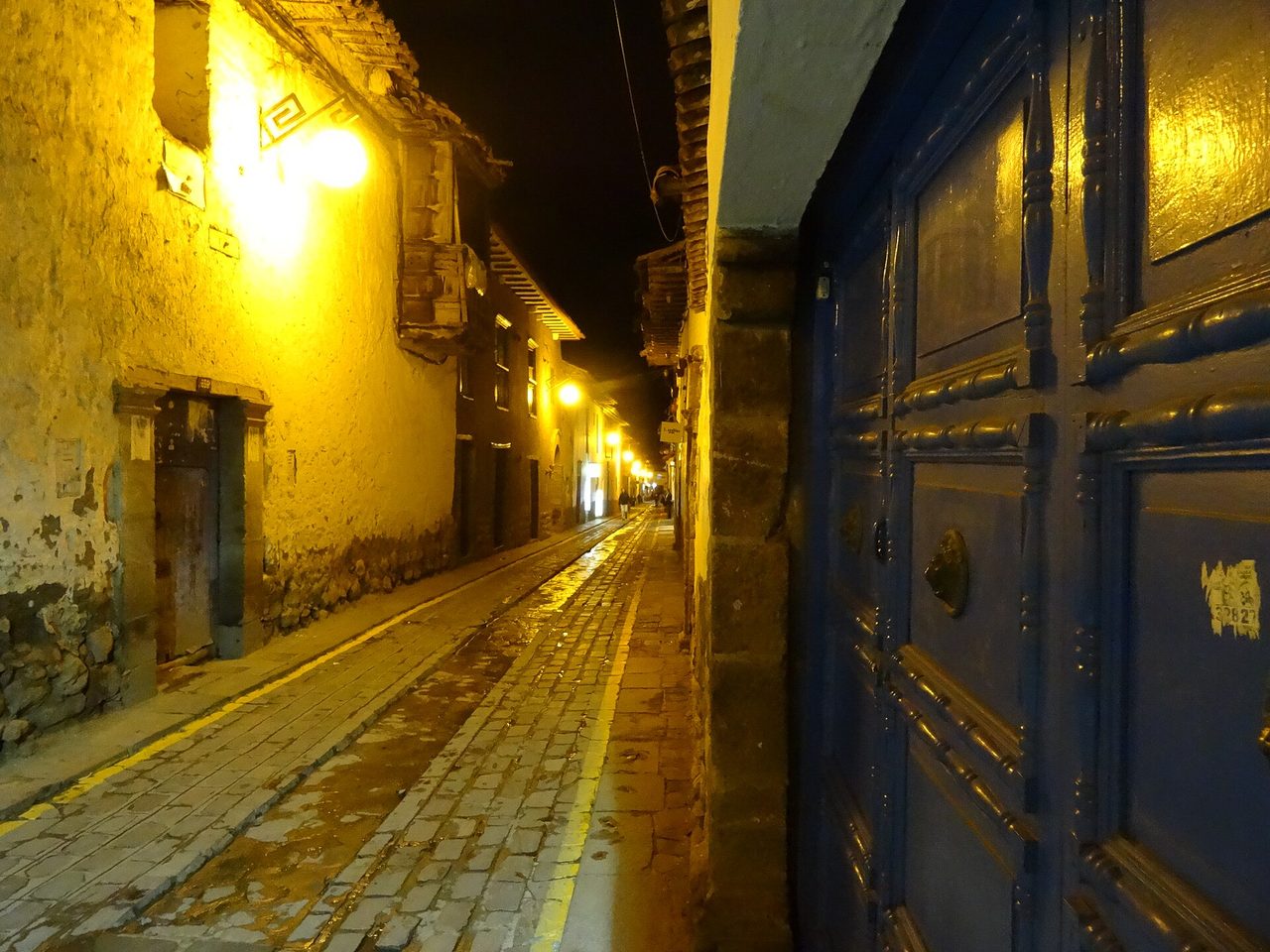 A narrow cobbled lane in Cusco lit by street lamps at night, running uphill towards San Blas past a heavy blue colonial door