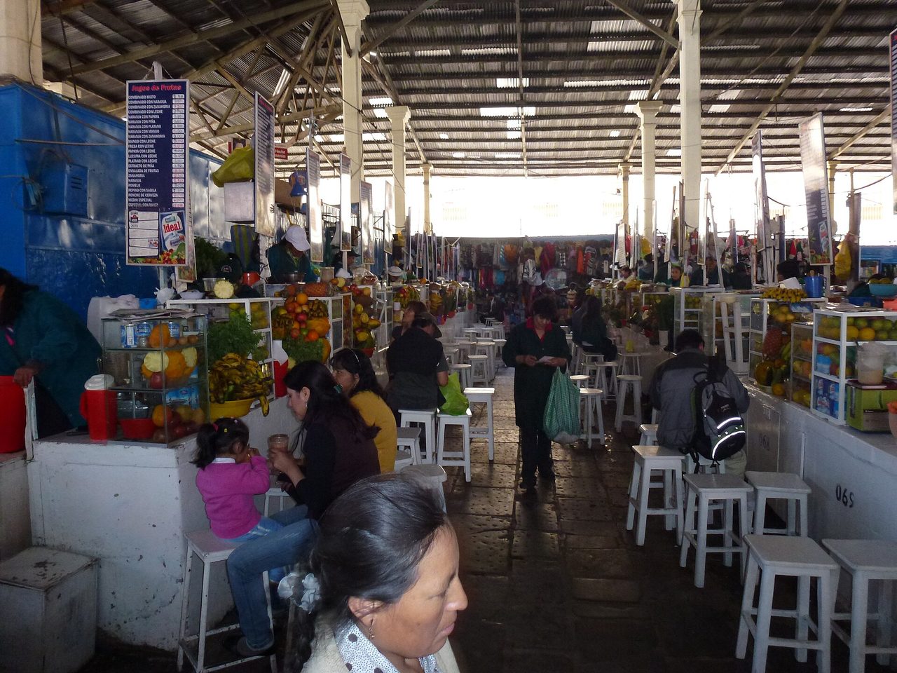 Numbered juice counters, fruit displays and white stools under the corrugated roof of the San Pedro market in Cusco, with shoppers passing between the aisles