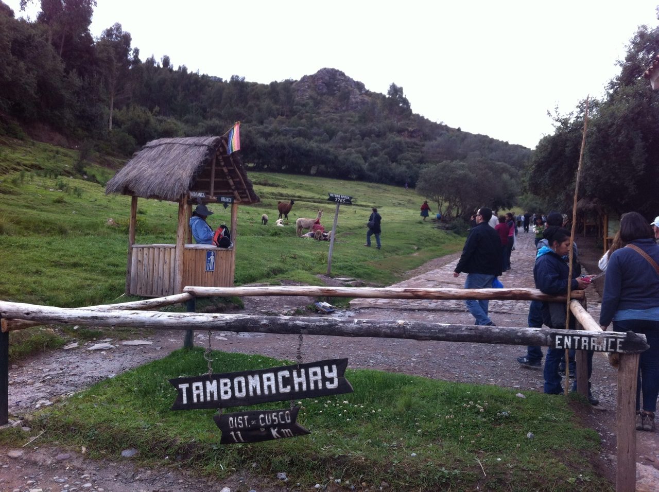 The wooden entrance barrier and hand-painted sign at Tambomachay, 11 km from Cusco, with llamas grazing on the hillside and visitors walking up the stone path