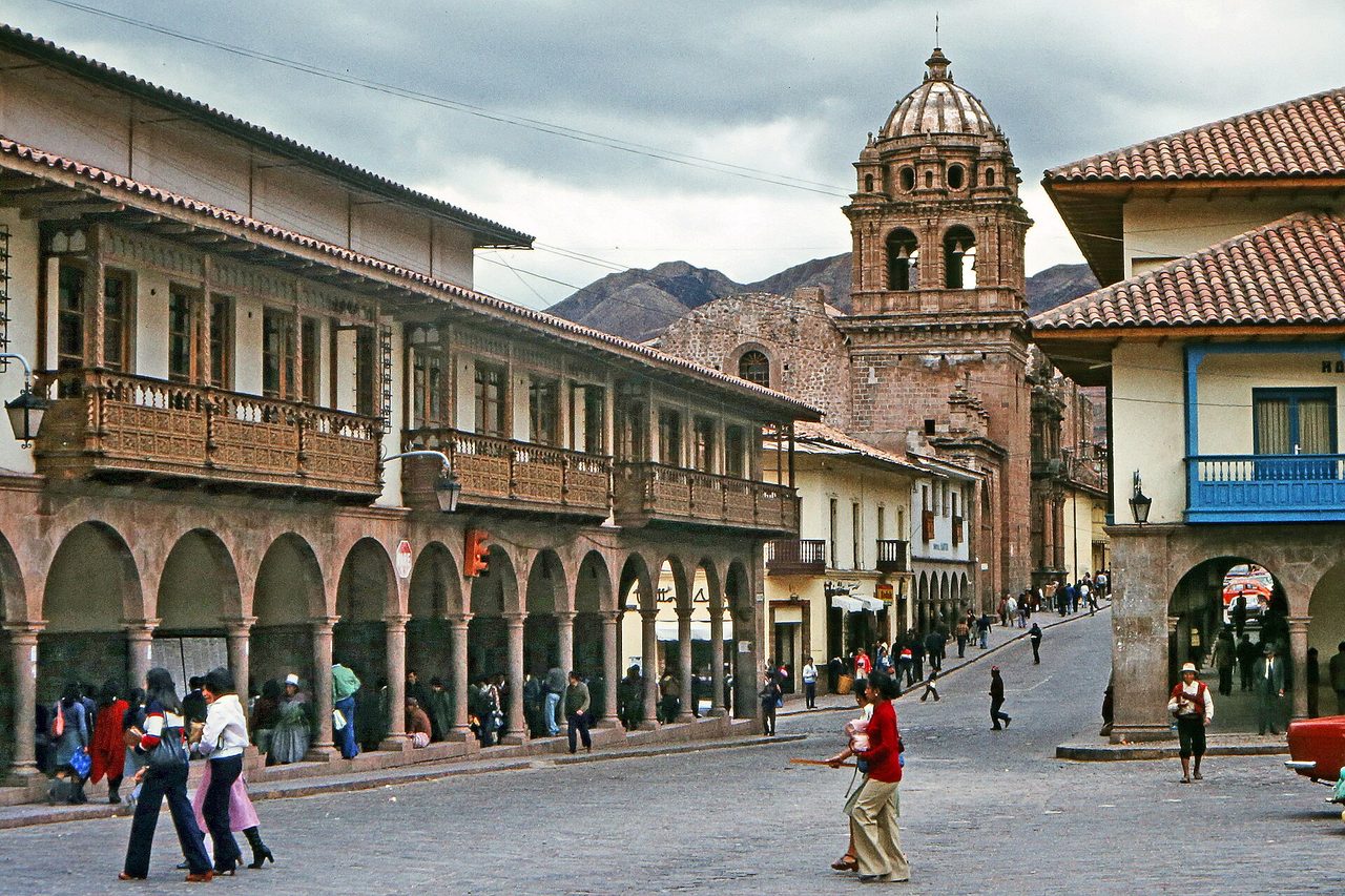 Cusco's Plaza de Armas under a grey overcast sky, with colonial arcades, carved wooden balconies and the bell tower of La Compania church