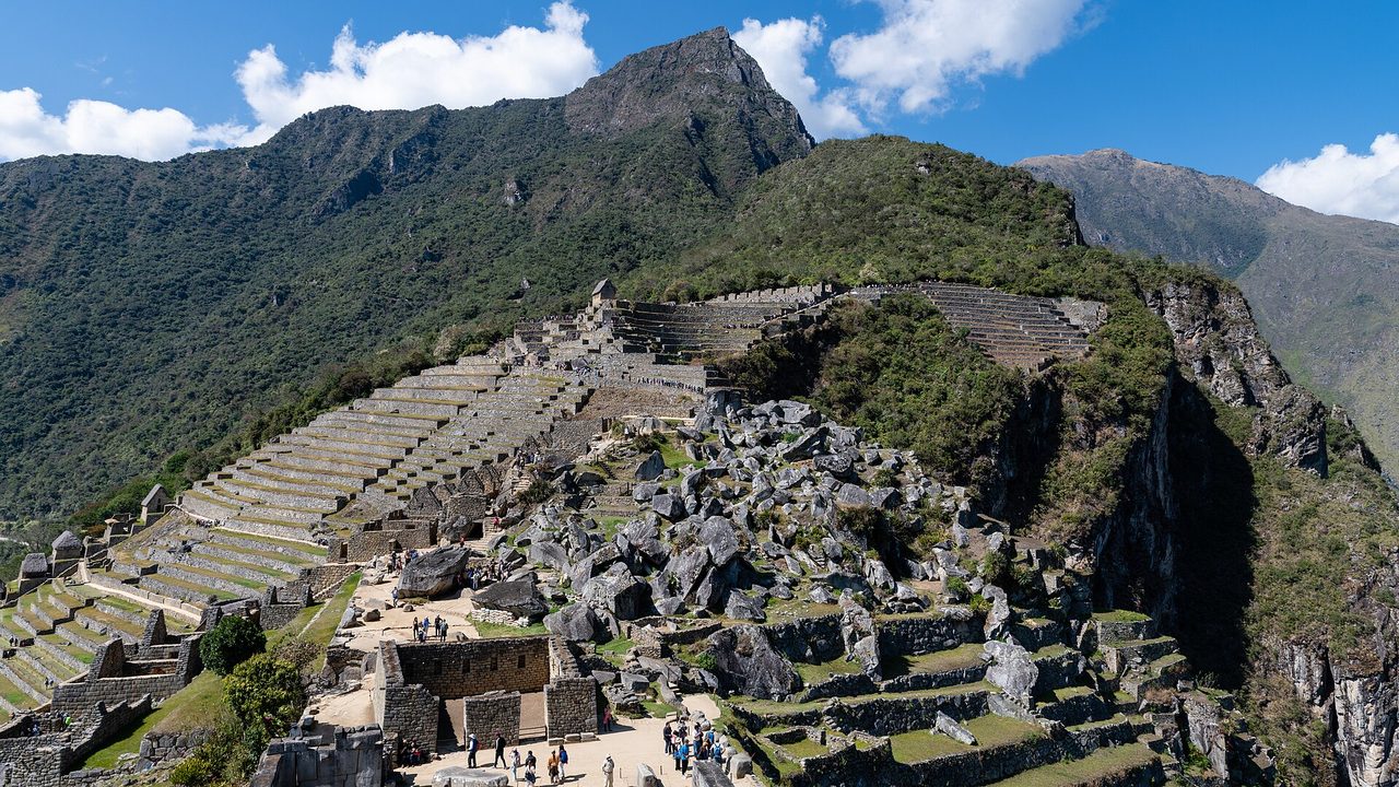 Machu Picchu's agricultural terraces and stone buildings under a clear blue sky, with small groups of visitors on the paths below