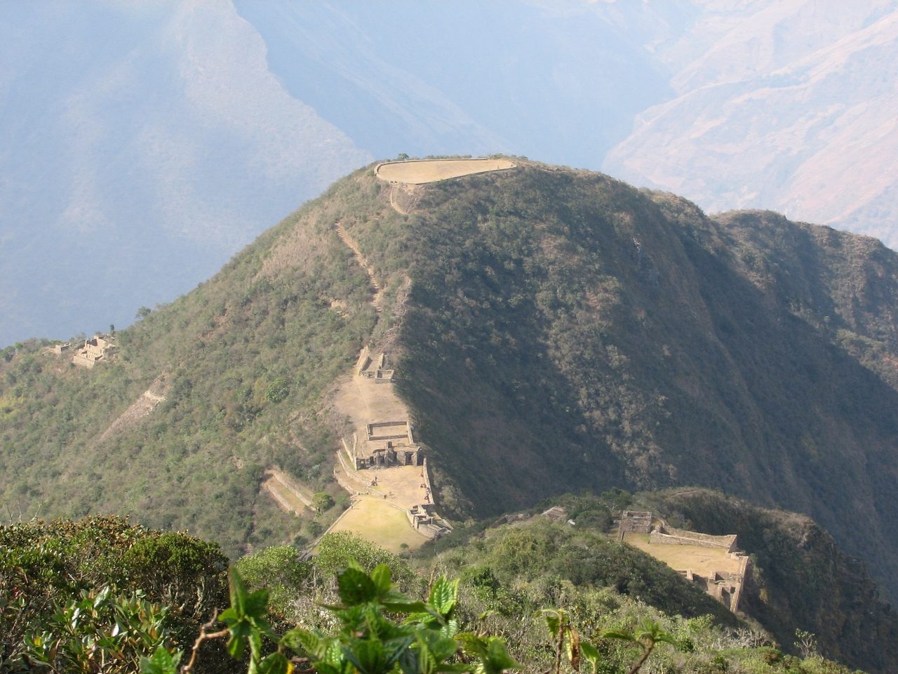 The ridge of Choquequirao seen from across the valley, with the flattened hilltop platform on top and stone buildings and terraces stepping down the spur