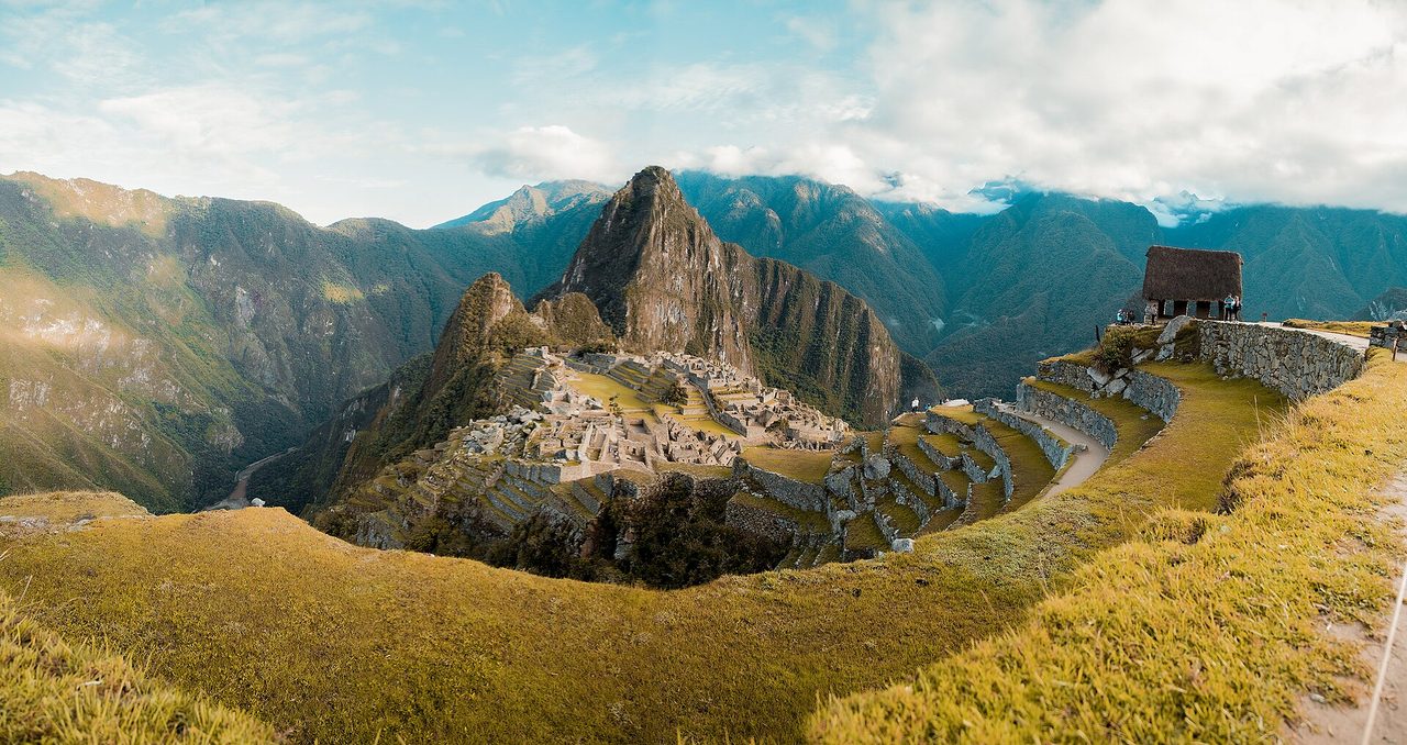 Panorama of the Machu Picchu citadel from the upper terraces, with the thatched guardian house on the right and Huayna Picchu rising behind the ruins