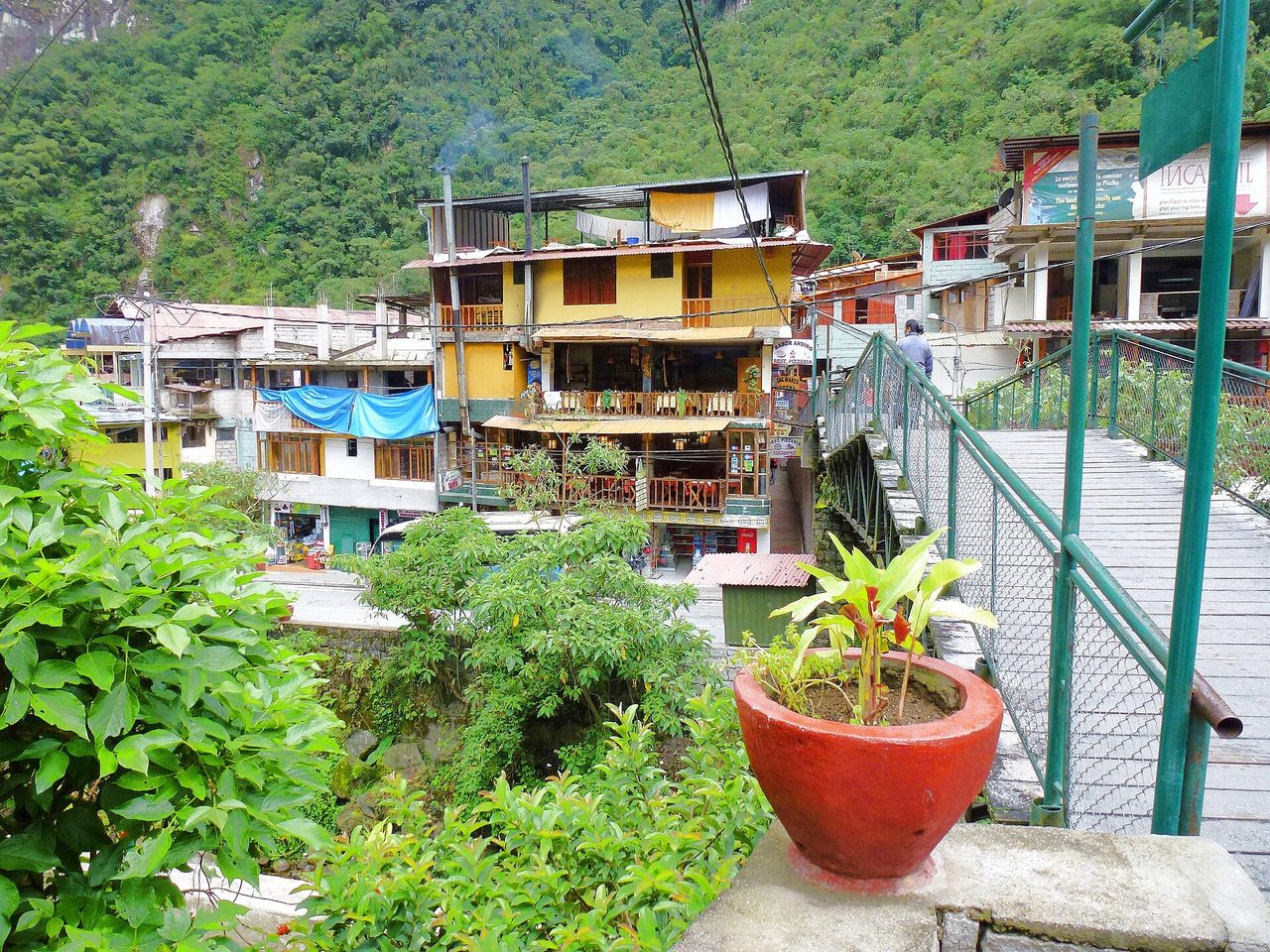 Hostels and restaurants stacked along a steep street in Aguas Calientes, with a footbridge over the river and forested cliffs behind