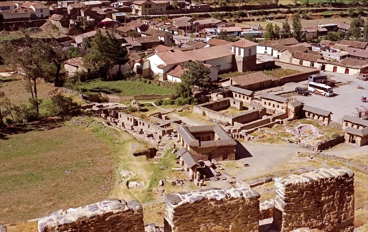 Ollantaytambo seen from above, with Inca ruins, village rooftops and tour buses parked beside the plaza