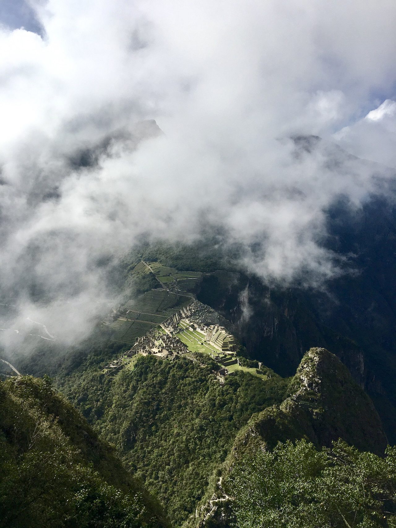 Machu Picchu seen from high above, the terraces and buildings small on their ridge as cloud pours across the surrounding mountains