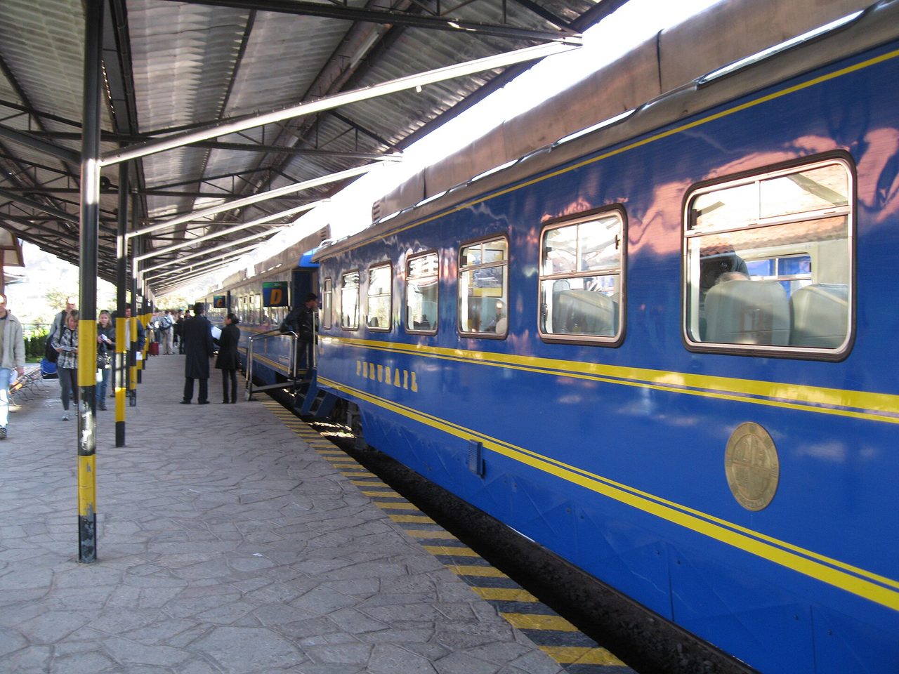 A blue and yellow PeruRail carriage waiting alongside a covered platform, with passengers boarding for the line to Aguas Calientes