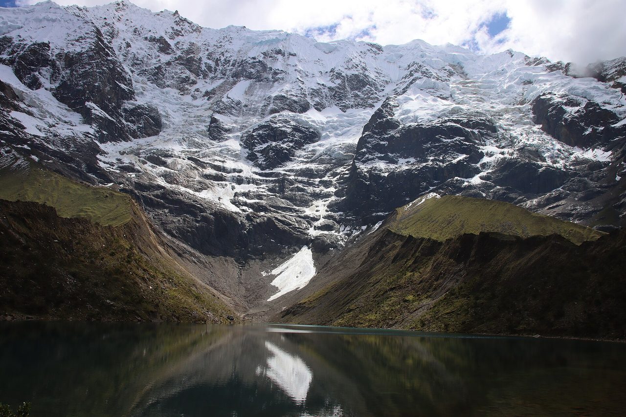 Humantay Lake under flat cloud, the water reading dark green rather than turquoise, below the heavily glaciated headwall that feeds it