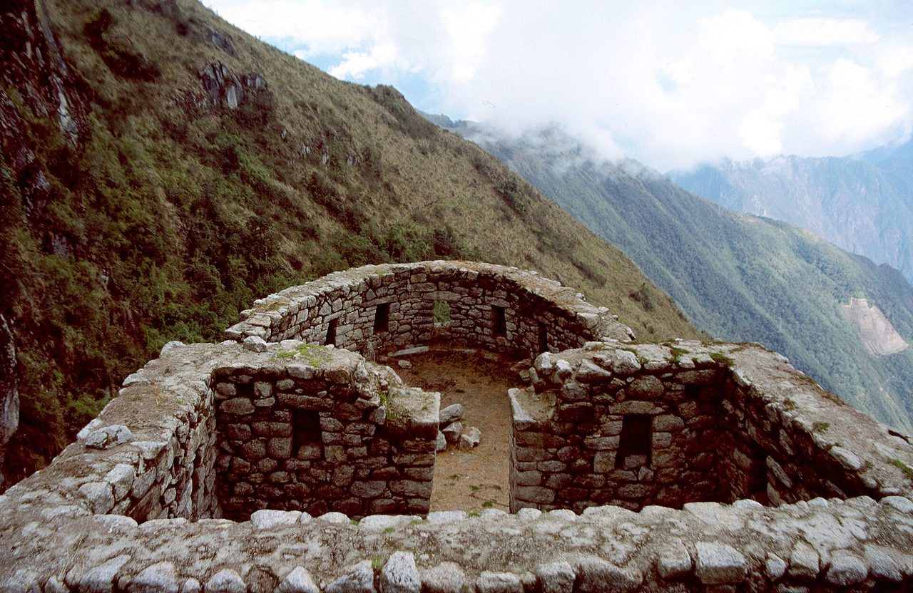 Curved stone walls and trapezoidal doorways of an Inca ruin on a ridge beside the Inca Trail, with cloud forest falling away behind