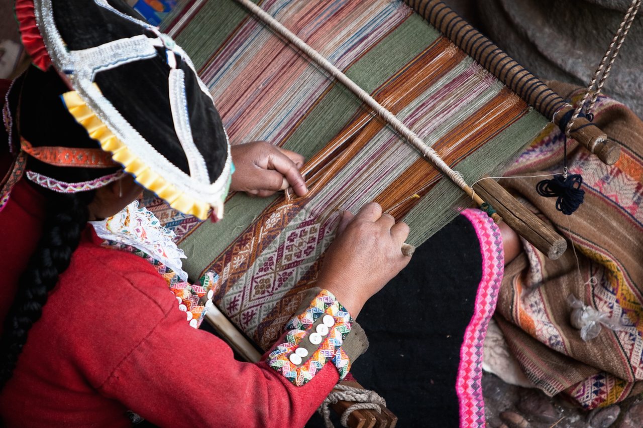 Close-up of an Andean woman weaving a patterned band on a backstrap loom, her hands working the warp threads