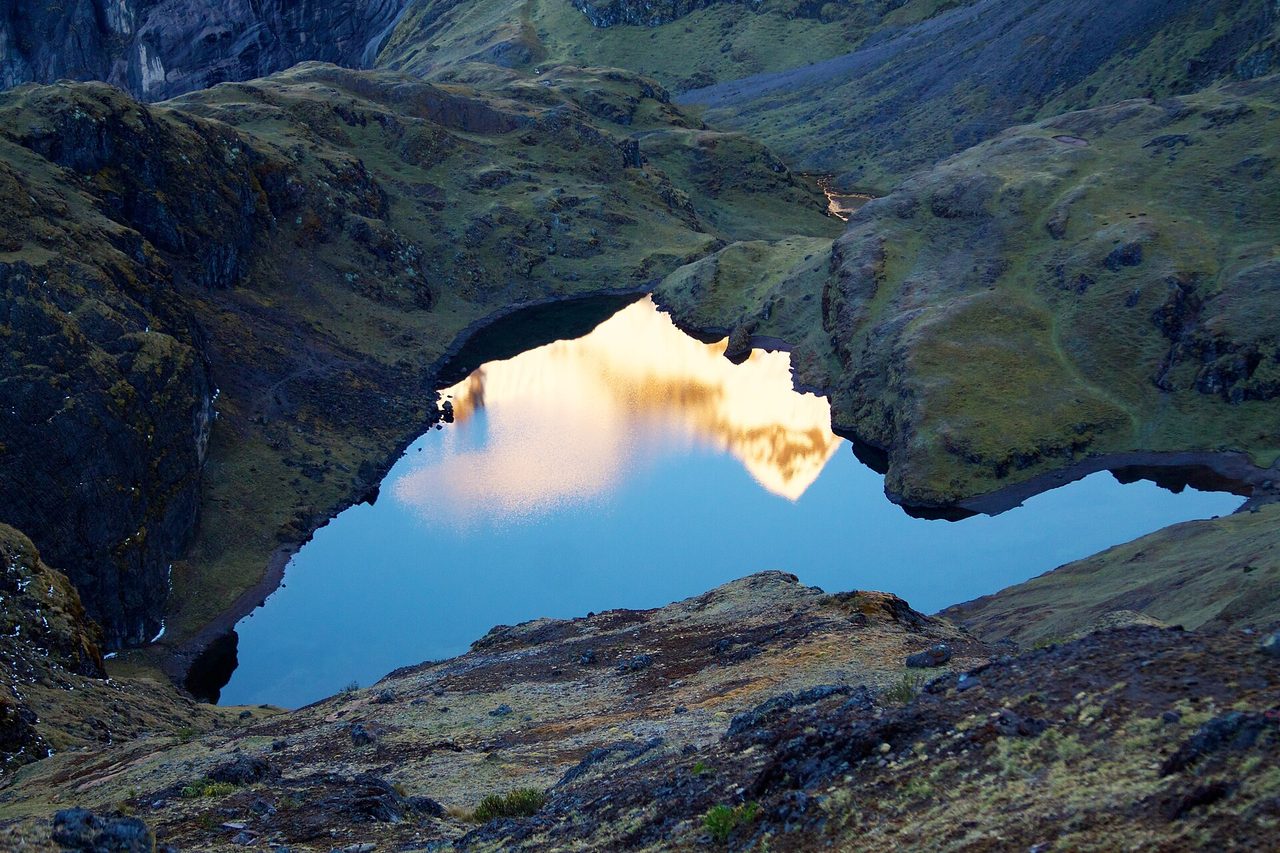 A still high-altitude lake on the Lares trek at dusk, reflecting a sunlit snow peak between treeless grassy slopes