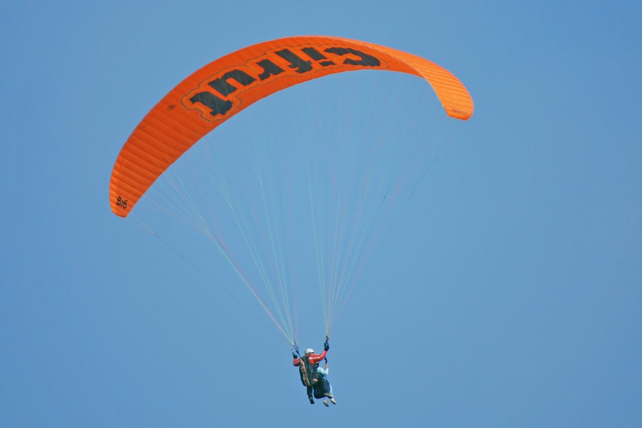 An orange tandem paraglider wing high over Miraflores against a clear sky