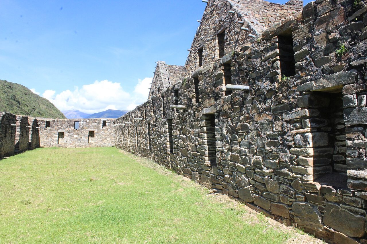 Restored Inca stone buildings with trapezoidal doorways lining a grassy plaza at Choquequirao, cloud-forest ridges behind