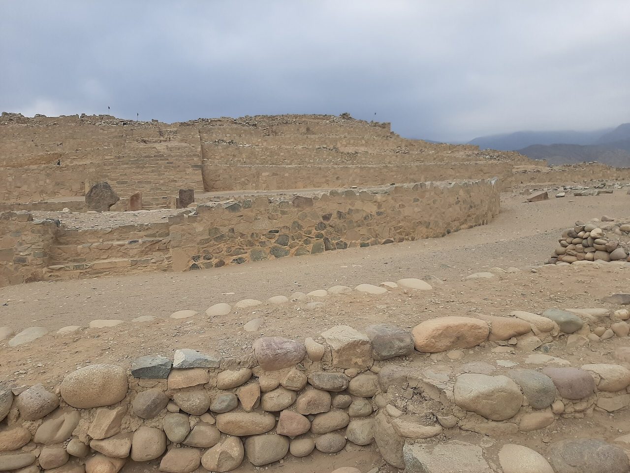 Stepped stone platform mound at Caral in the Supe valley, with river-cobble retaining walls in the foreground under a grey desert sky