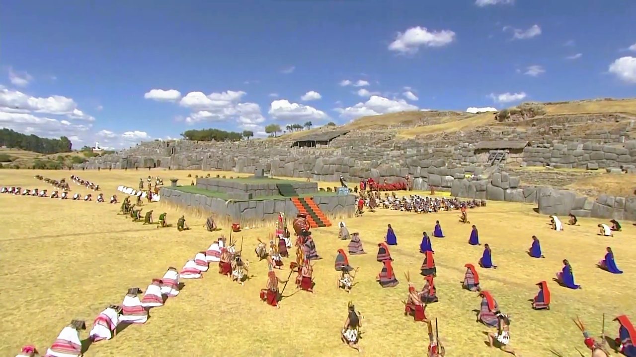 Hundreds of costumed performers in coloured cloaks kneeling and standing on the yellow grass esplanade at Sacsayhuaman during the Inti Raymi ceremony, with the zigzag Inca walls behind
