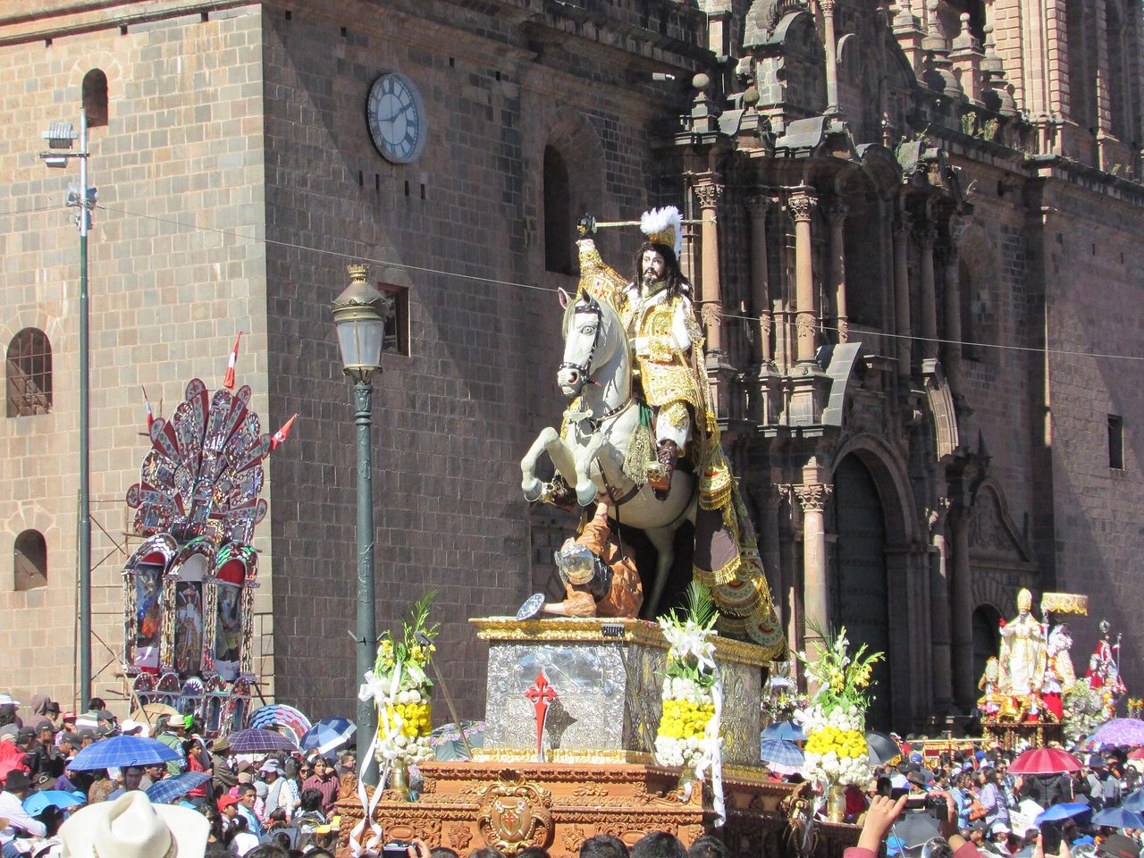 The image of Santiago on a white horse, mounted on a silver and gilt litter, carried past the facade of Cusco Cathedral through a crowd of umbrellas during the Corpus Christi procession