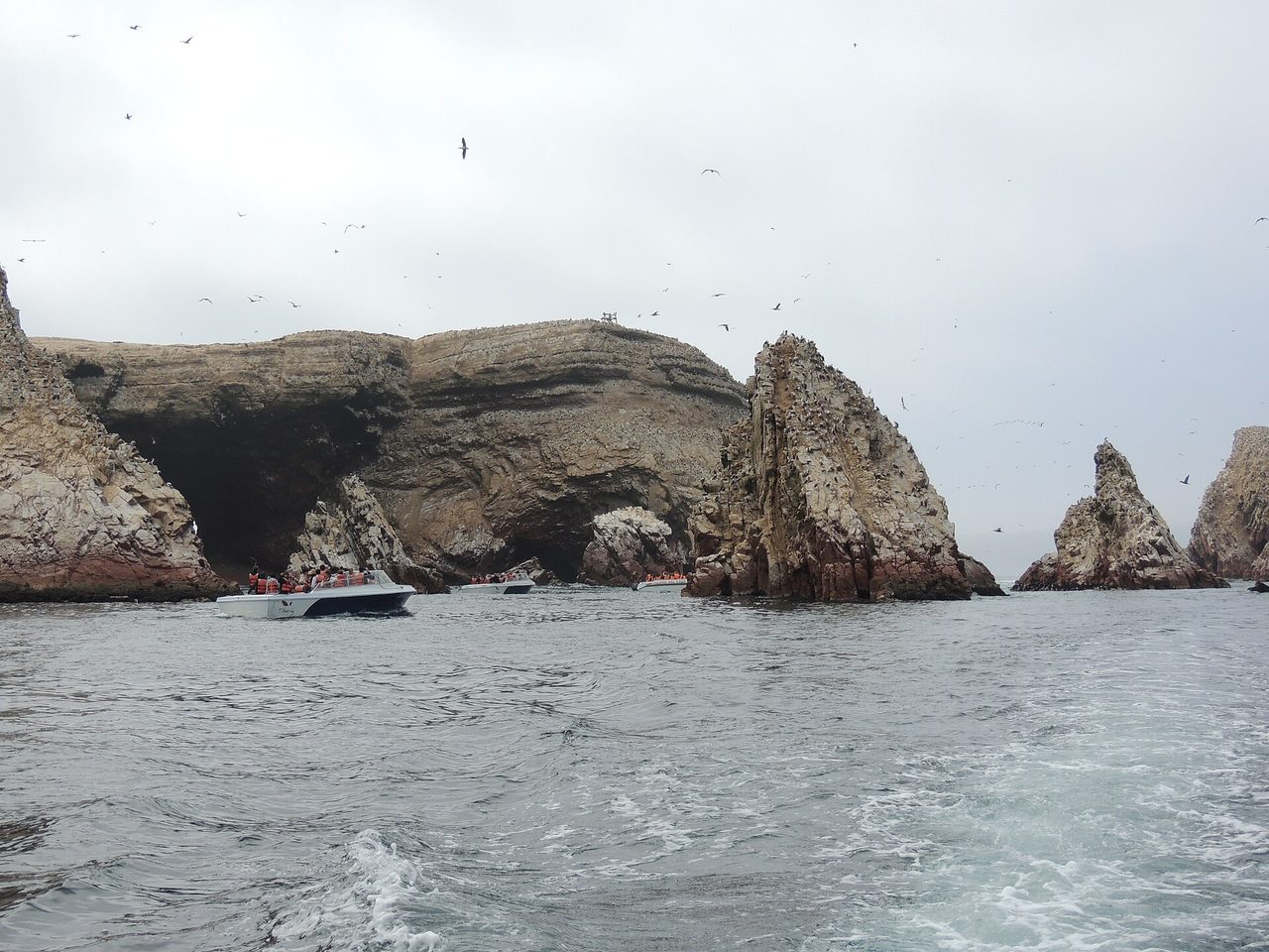 Tour boats beneath the guano-streaked cliffs of the Ballestas Islands off Paracas under a flat grey sky, with seabirds wheeling overhead
