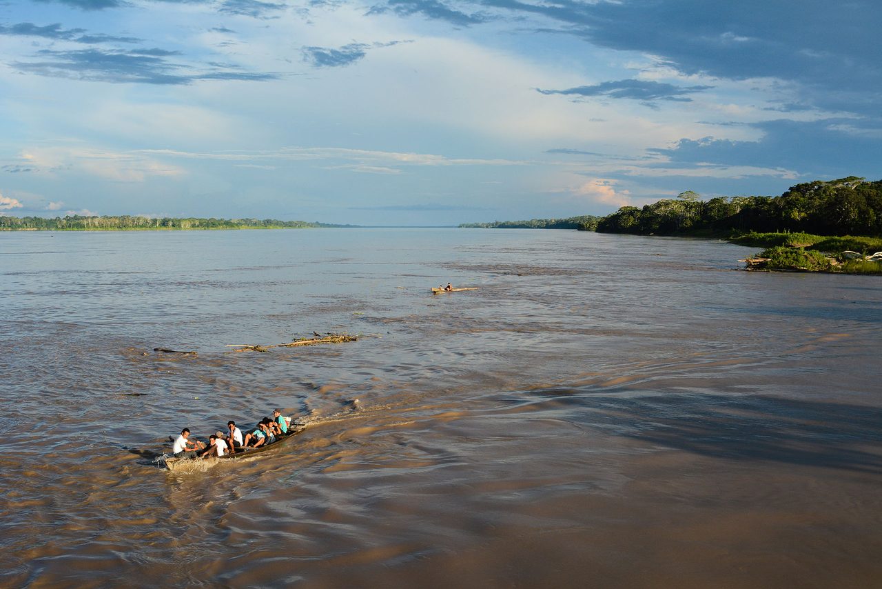 Dugout canoes carrying passengers across a broad brown stretch of the Amazon river with dense rainforest along both banks