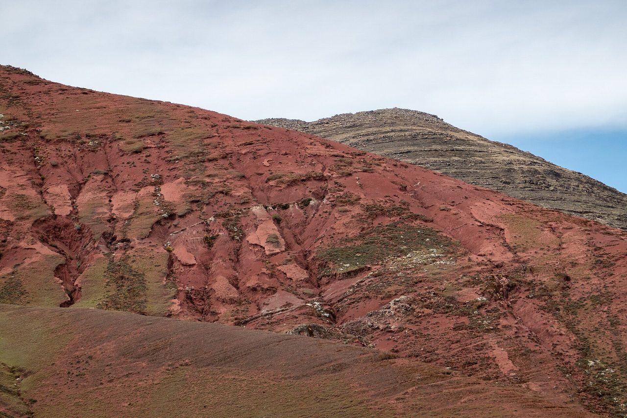 A deep red mineral slope in the Palccoyo rainbow range near Checacupe, with layered grey rock capping the ridge above