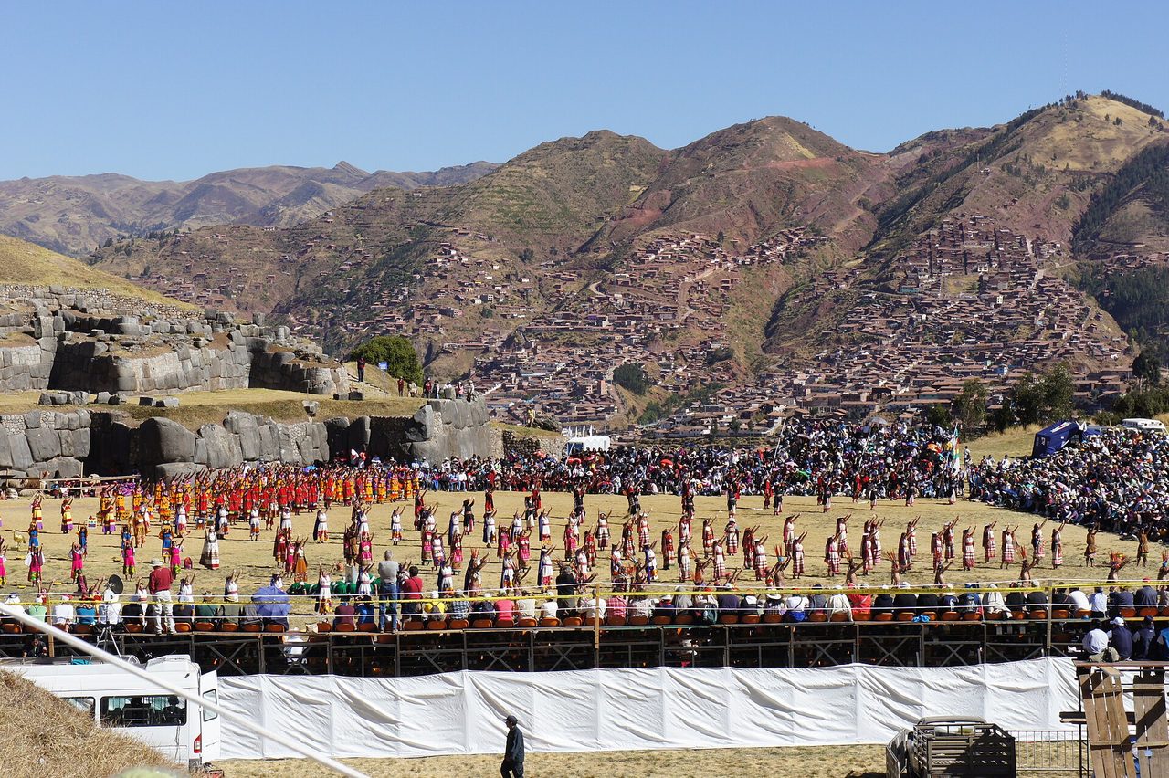 Hundreds of costumed performers on the Sacsayhuaman esplanade during Inti Raymi, with temporary grandstands in the foreground and the Inca ramparts behind