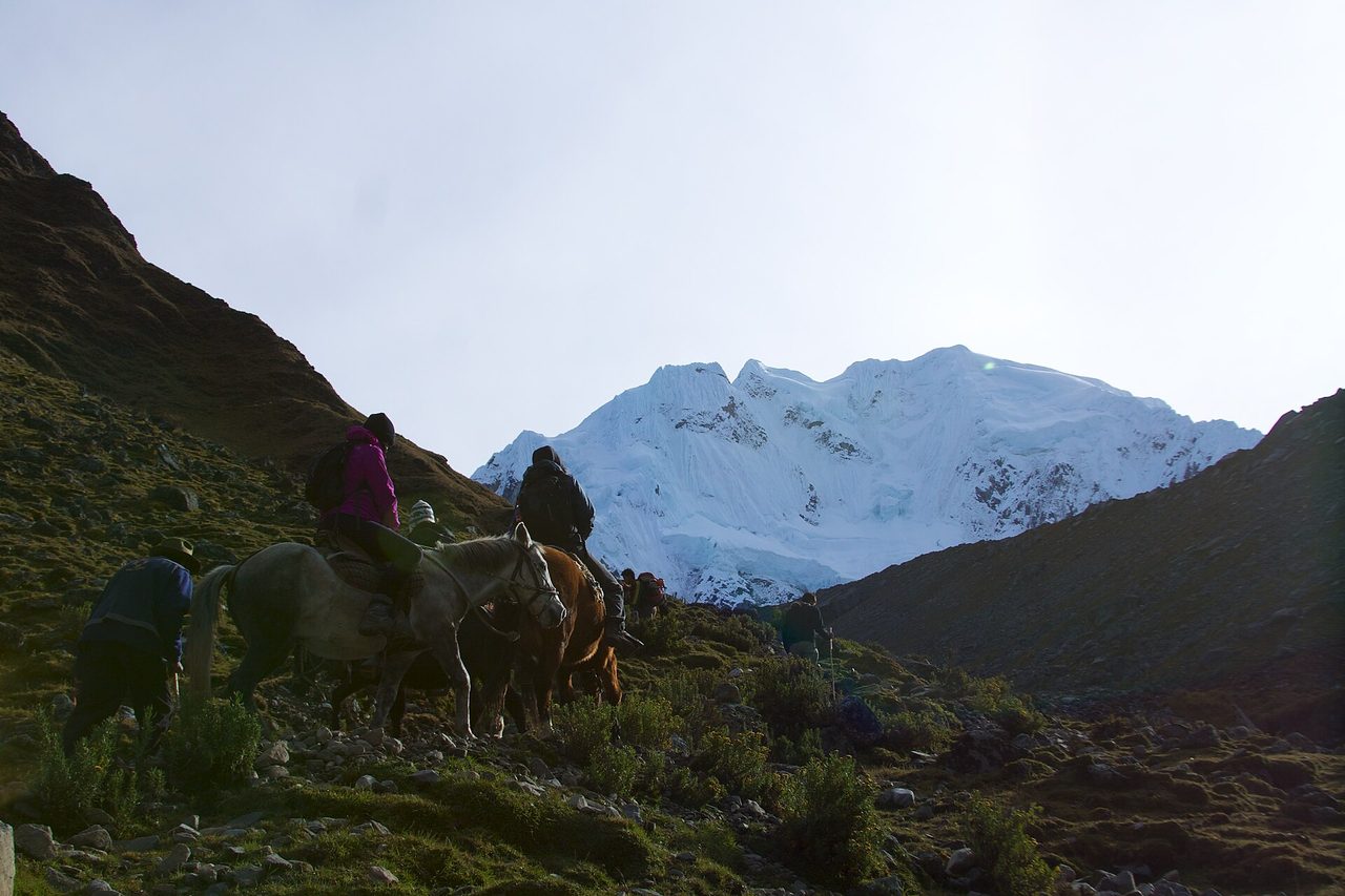 Riders and pack horses on a stony track at dawn below the glaciated face of Salkantay