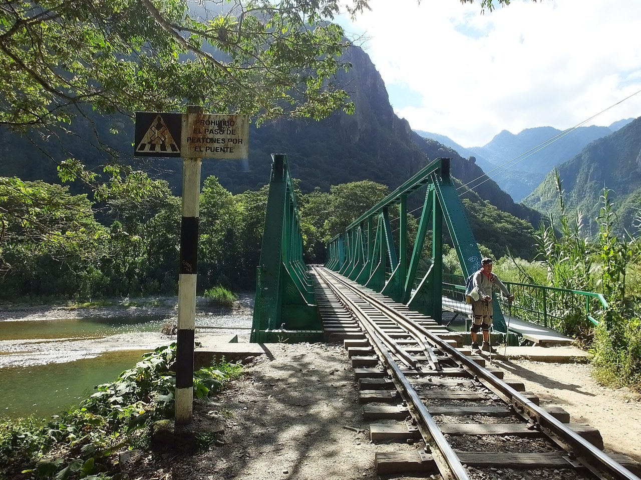 A hiker beside a green steel railway bridge over the Urubamba river on the track walk into Aguas Calientes