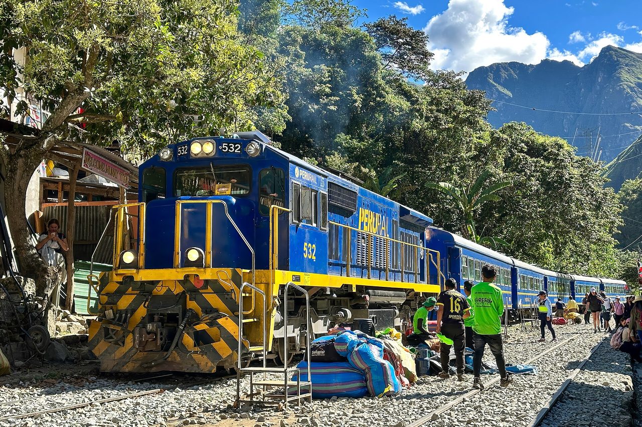 A blue PeruRail train stopped on the track with passengers loading gear from the ballast, since there is no platform on this line