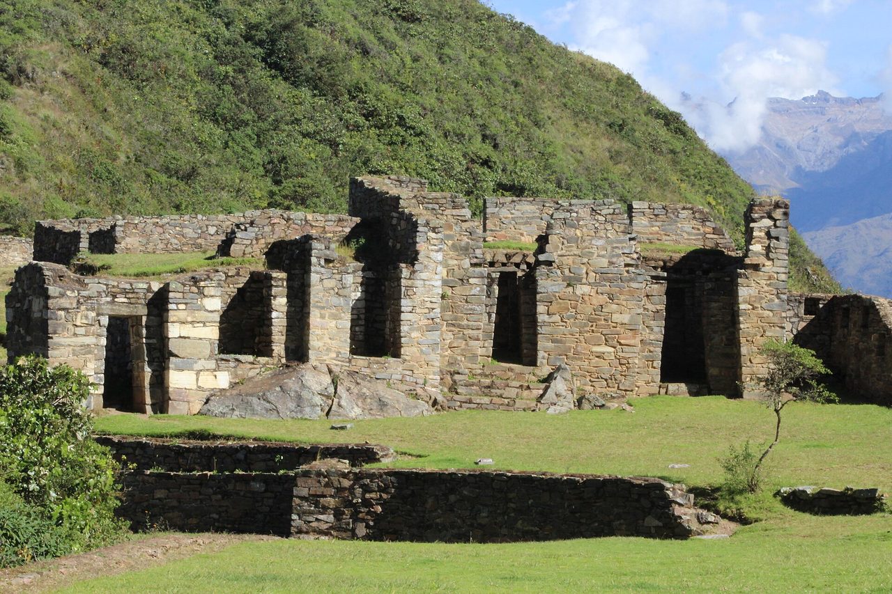 A row of Inca stone buildings with trapezoidal doorways at Choquequirao, with green ridges and a snow-capped peak behind