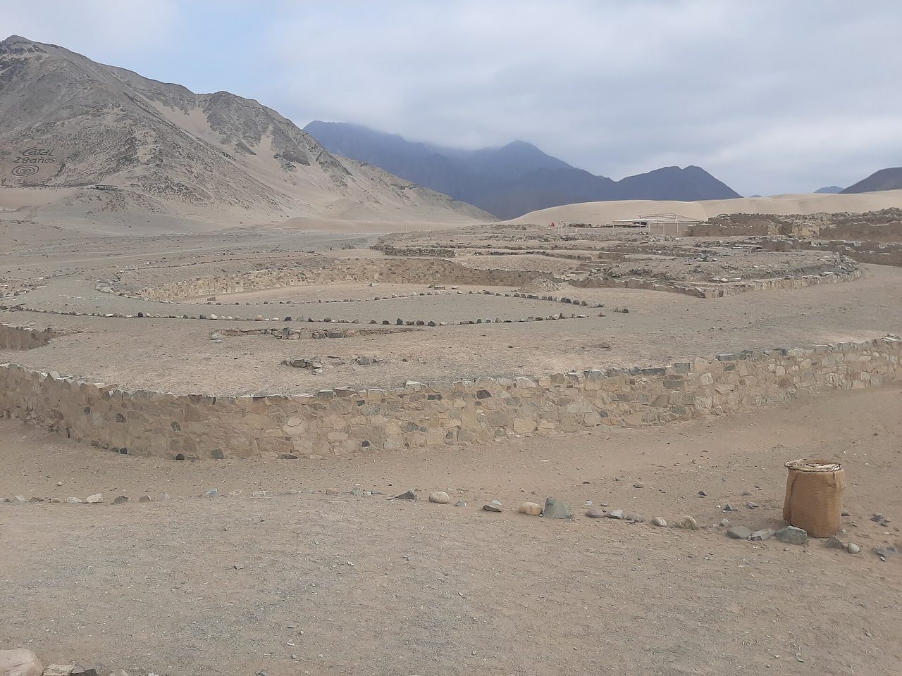 Low stone walls and a sunken circular plaza at the desert site of Caral in Peru's Supe valley, with arid hills behind