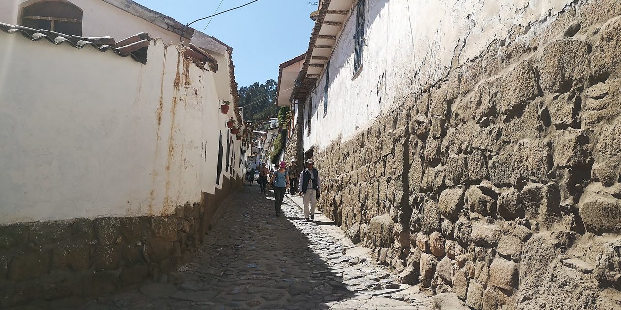 The narrow cobbled lane of Calle Tandapata climbing through San Blas in Cusco between a whitewashed wall and a massive Inca stone wall, with a few walkers coming down it