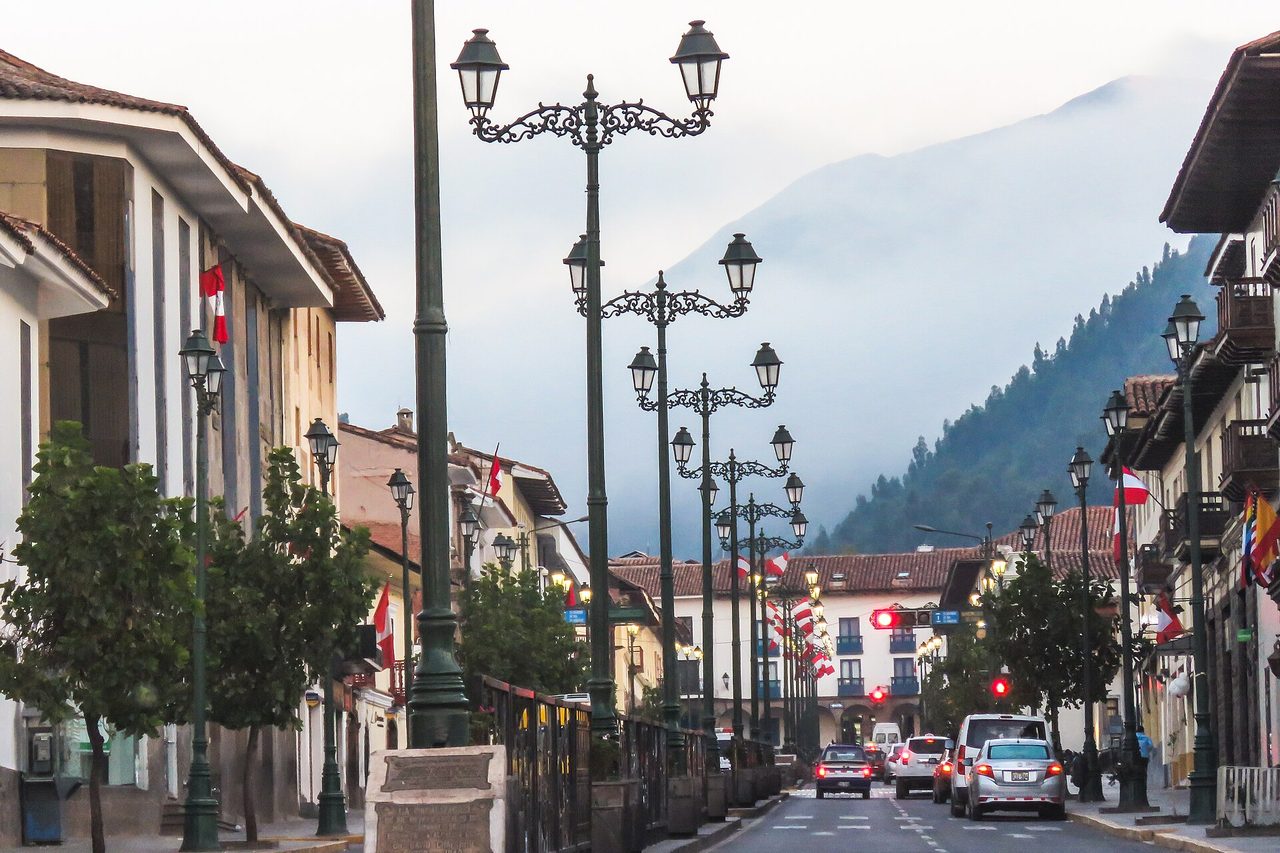 A wide, flat, tree-lined avenue in central Cusco with ornate iron street lamps, cars at a traffic light and Peruvian and Cusco flags on the balconies, the valley hills rising behind