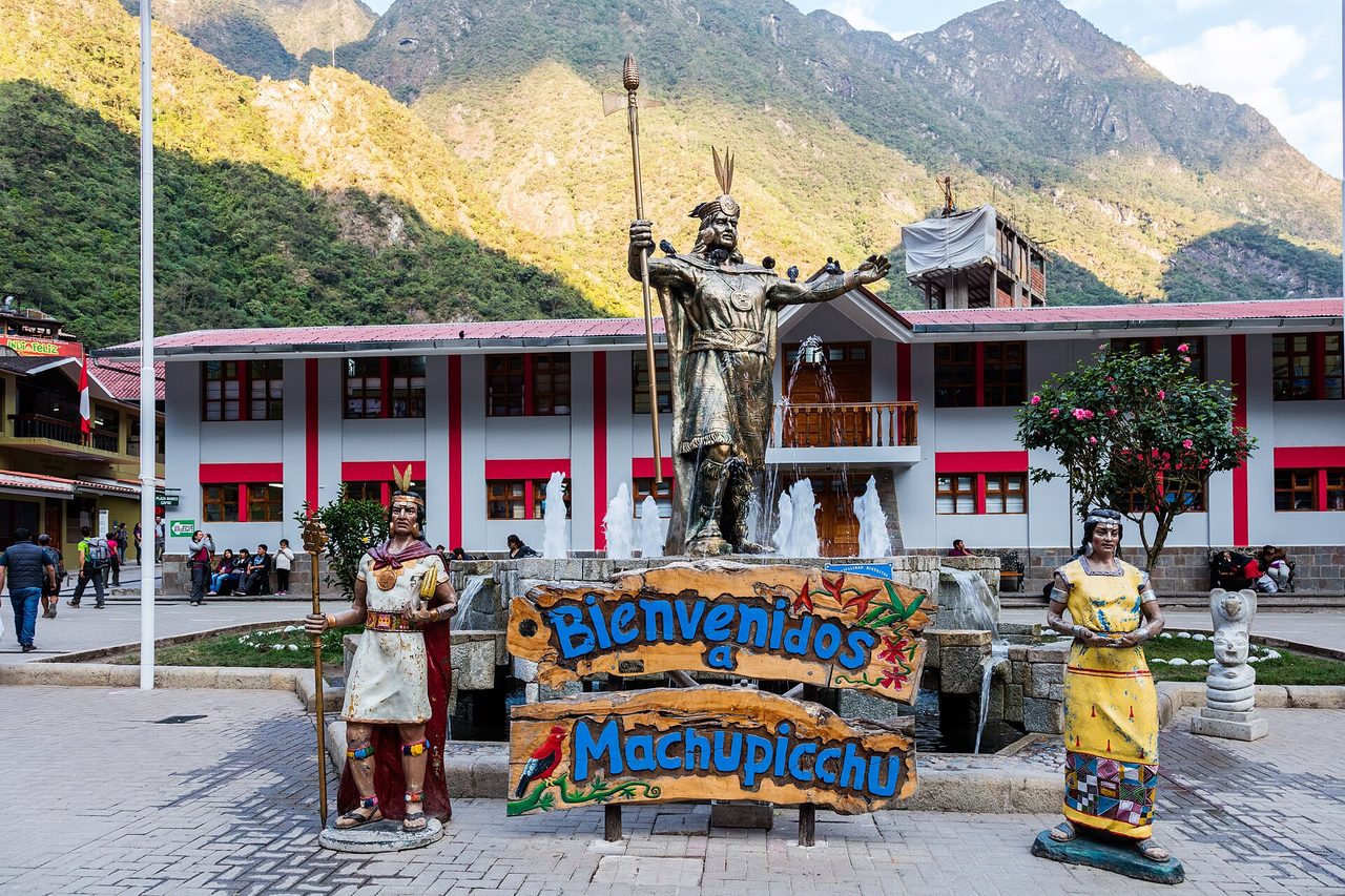 The welcome sign, fountain and Inca statue in the main square of Aguas Calientes, with forested peaks behind