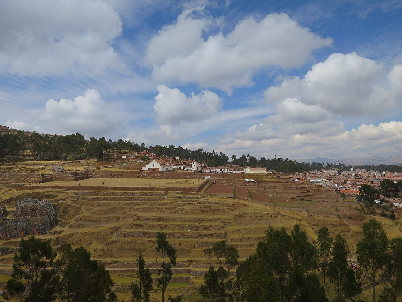 Wide view of the Chinchero archaeological site: the white church above a broad Inca esplanade, terraces below and the modern town to the right