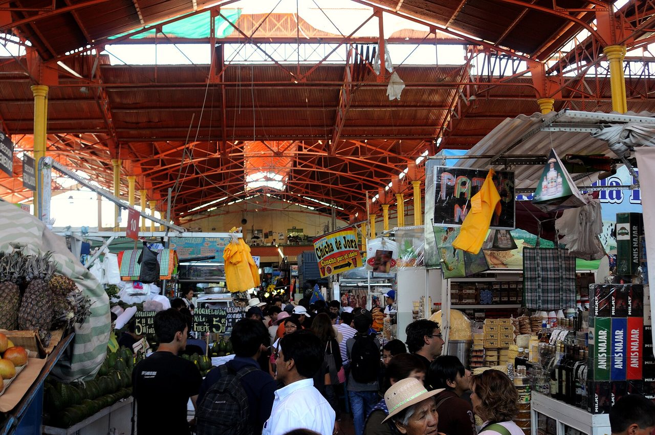 Crowded aisles and produce stalls under the iron roof of San Camilo market in Arequipa, with prices chalked up in soles per kilo