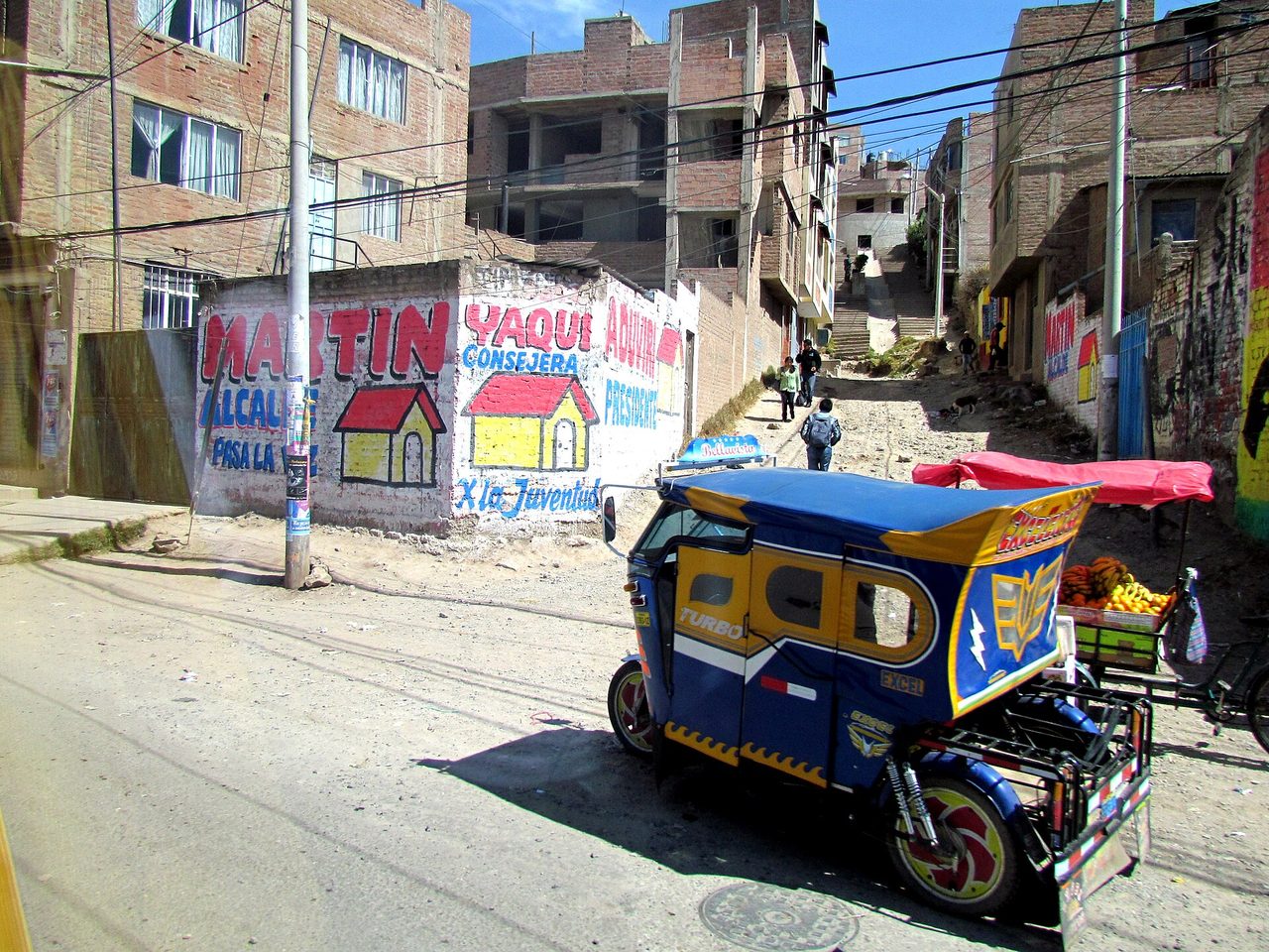 A blue and yellow mototaxi parked on a steep unpaved street in Puno beside a fruit cart