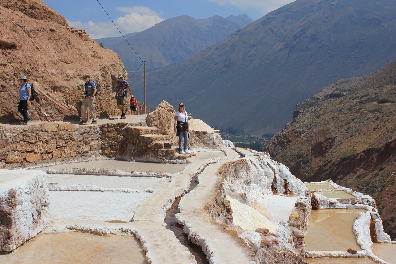 Visitors on the narrow walkway above the Maras salt pans, with the shallow pools and their earth dividers below and the valley beyond