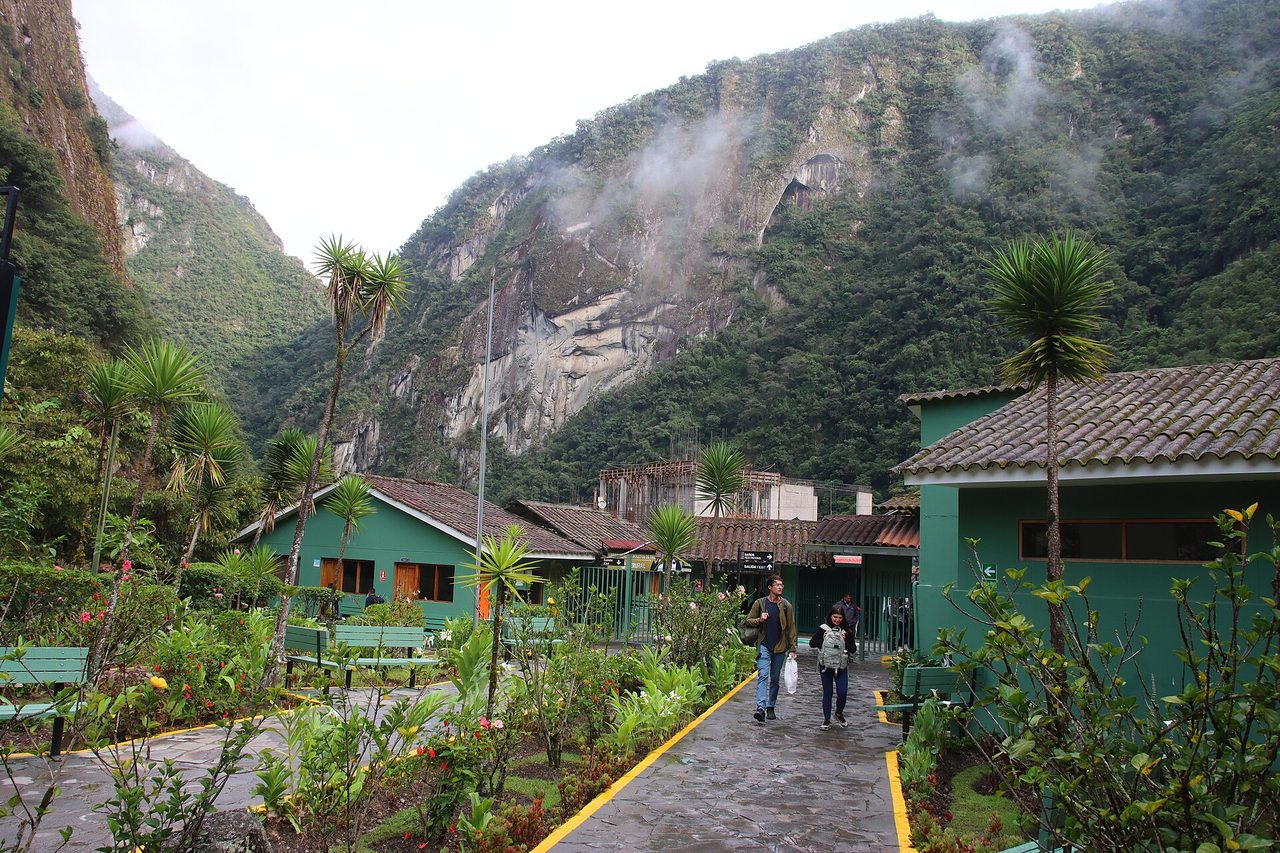Passengers walking out of the green station buildings at Aguas Calientes with cloud forest cliffs behind