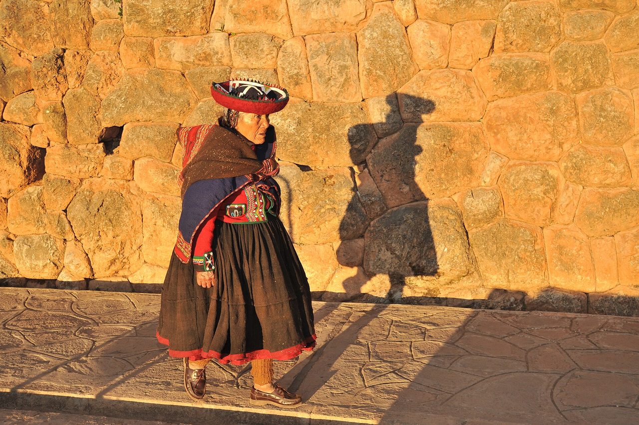 A woman in the flat red hat and layered skirts typical of Chinchero walking past a massive Inca stone wall in late-afternoon light