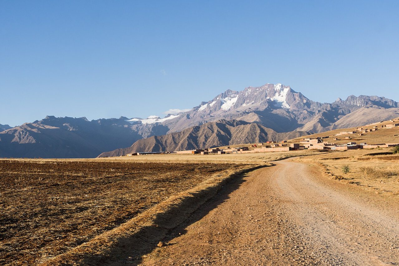 A glaciated Andean peak rising behind a plateau village in the Cusco region, seen across harvested fields from a dirt road