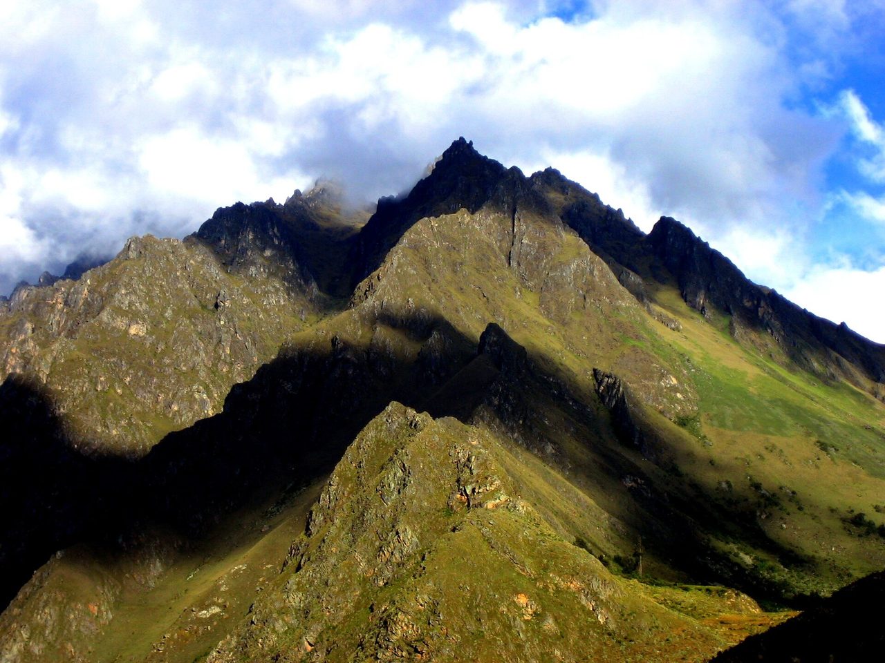 Steep grass-and-rock ridges in the Andes near the Urubamba valley, with cloud spilling over the summit