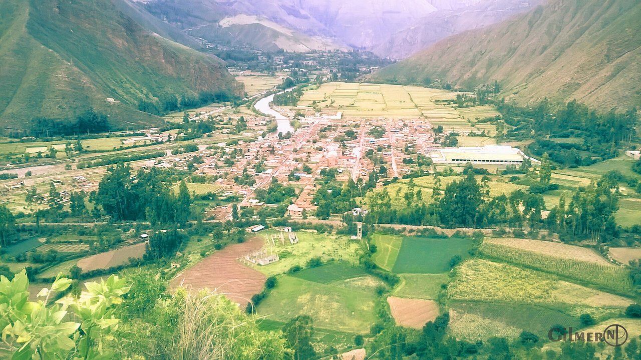 A valley-floor town in Urubamba province seen from the hillside above, with the river and a patchwork of farm plots filling the flat ground between steep ridges