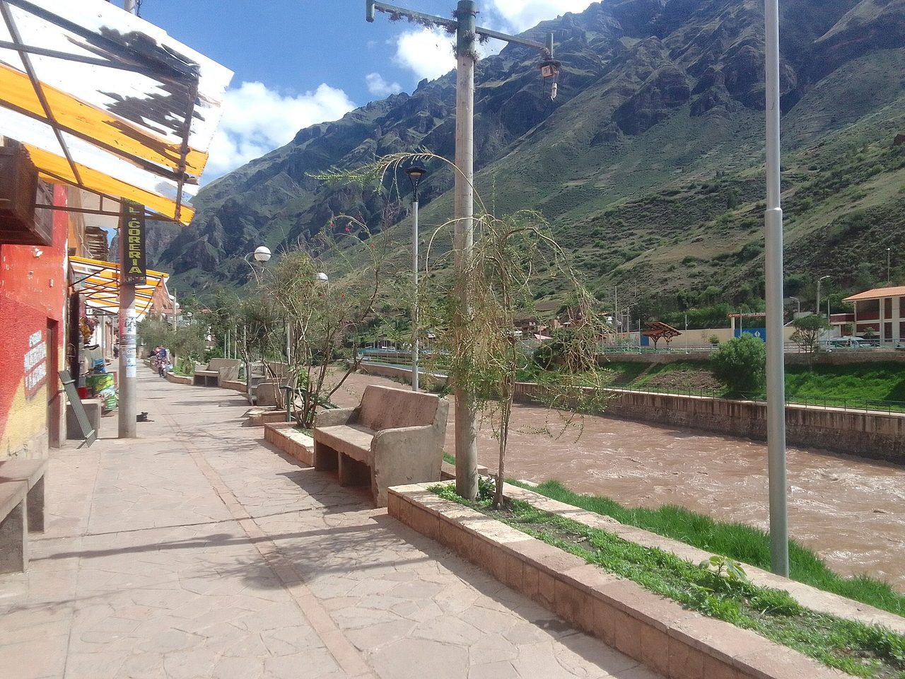 A riverside street in a small Sacred Valley town, with the Urubamba river running high and brown alongside the pavement